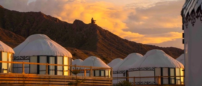 Several yurts on wooden platforms against mountains at sunset, with a person sitting on a distant peak.