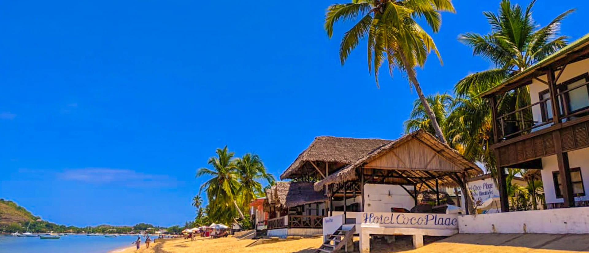 Sunny beach in Madagascar with Hotel Coco Plage, palm trees, and people strolling by the ocean.