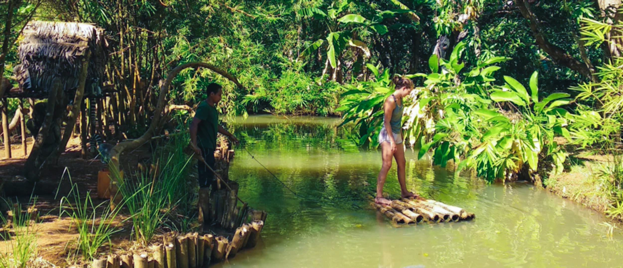 A woman stands on a bamboo raft in a green river, guided by a man on the bank.