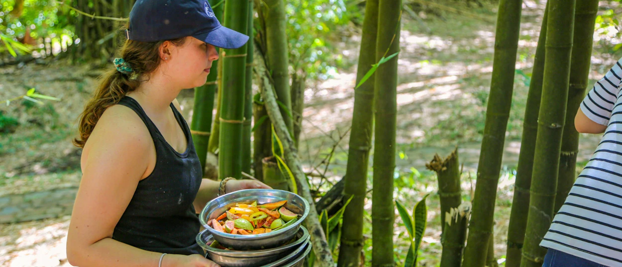 A woman in a blue cap carries stacked bowls of chopped fruit and vegetables in a bamboo forest.