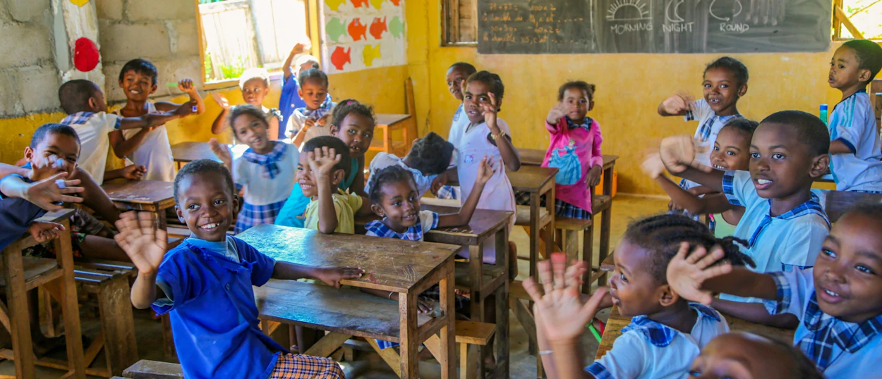 Joyful students in a bright classroom in Madagascar smiling and waving from their desks.