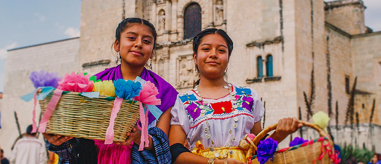 Two young women in traditional Oaxacan clothing hold baskets of colorful crafts outside a cathedral.
