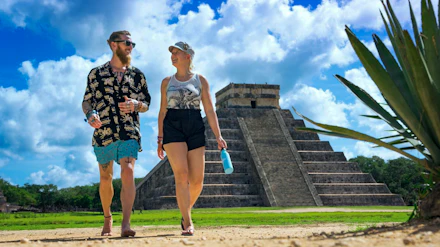 A man and woman stroll past the impressive Chichen Itza pyramid in Mexico under a bright sky.