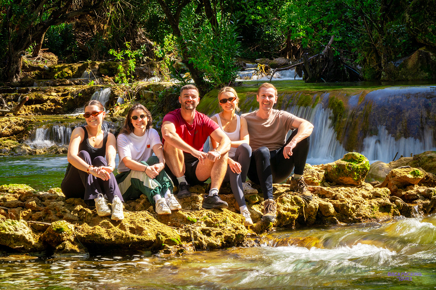 A group of travellers sits on the rocks in front of a small waterfall at Akchour.