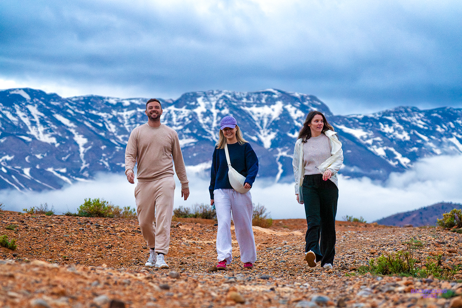 Three travellers walk along a mountain path above the clouds with snowy Atlas peaks behind.