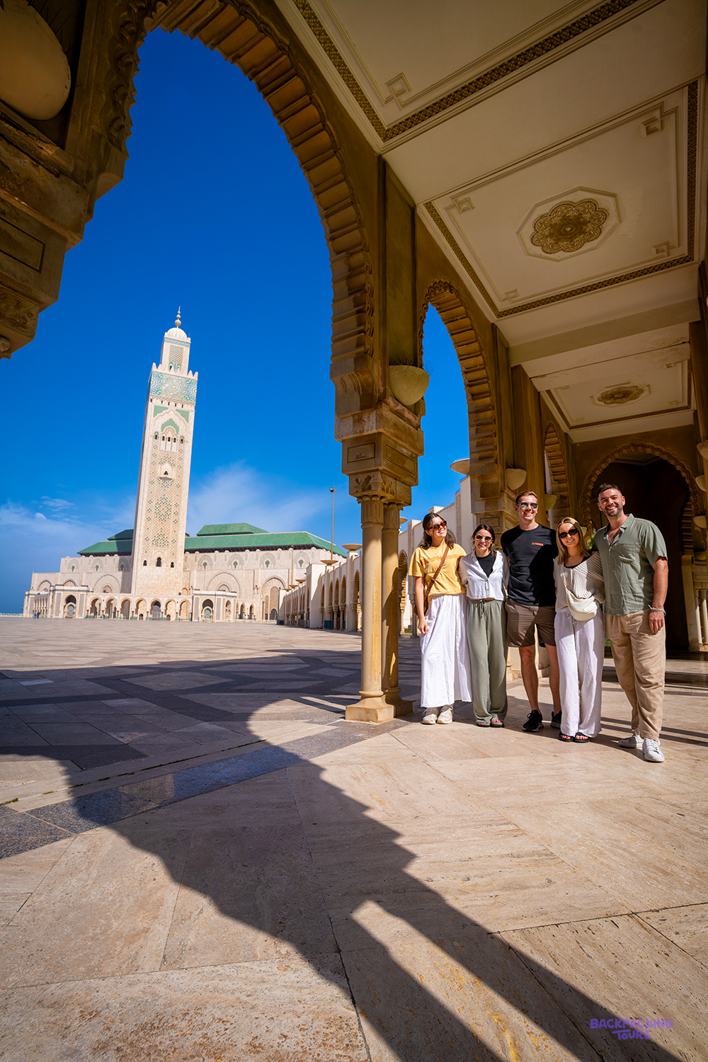 Five travellers smile beneath a carved archway with the tall minaret of the Hassan II Mosque behind.