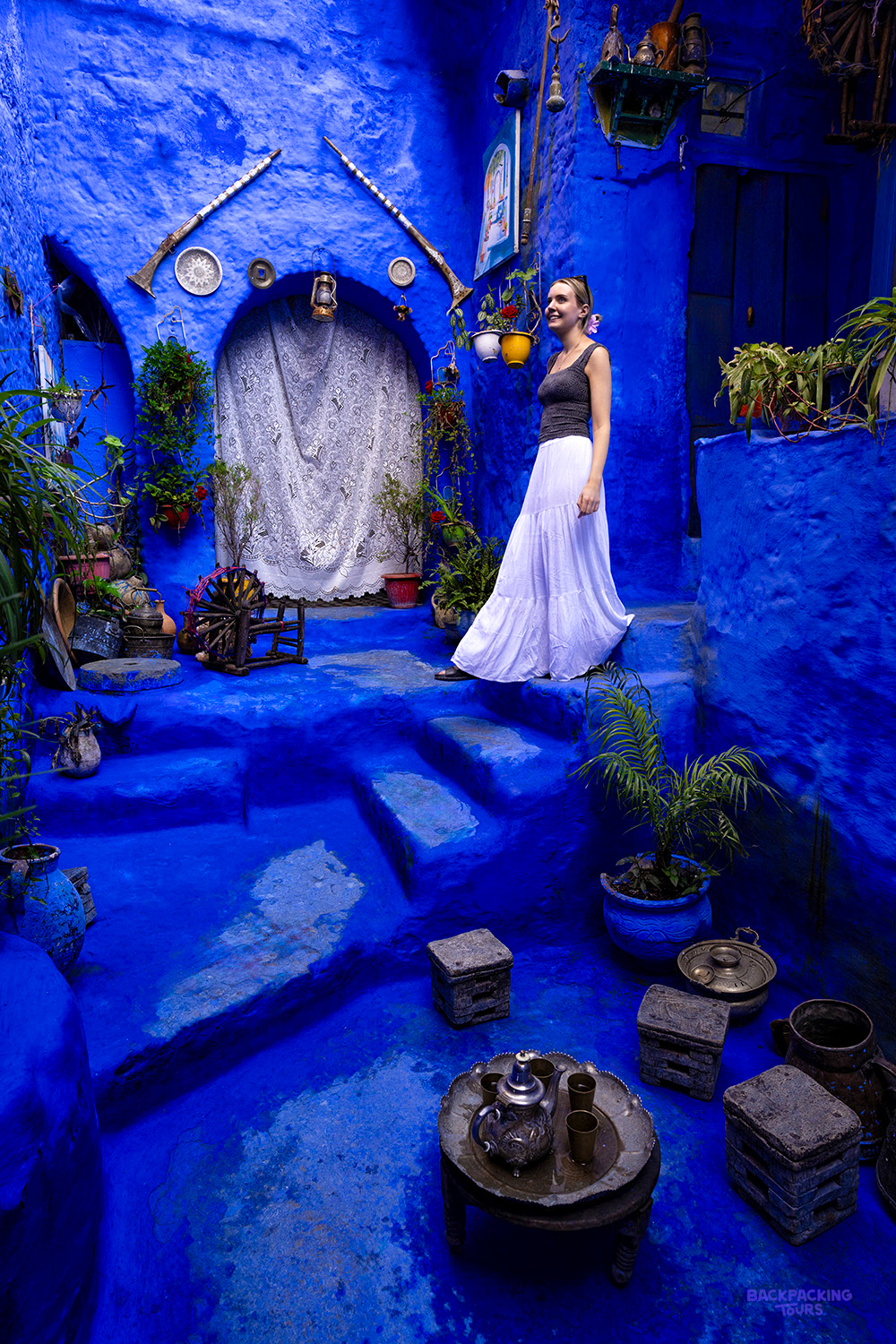 A traveller in a long white skirt stands on a bright blue stairway lined with plants in Chefchaouen.