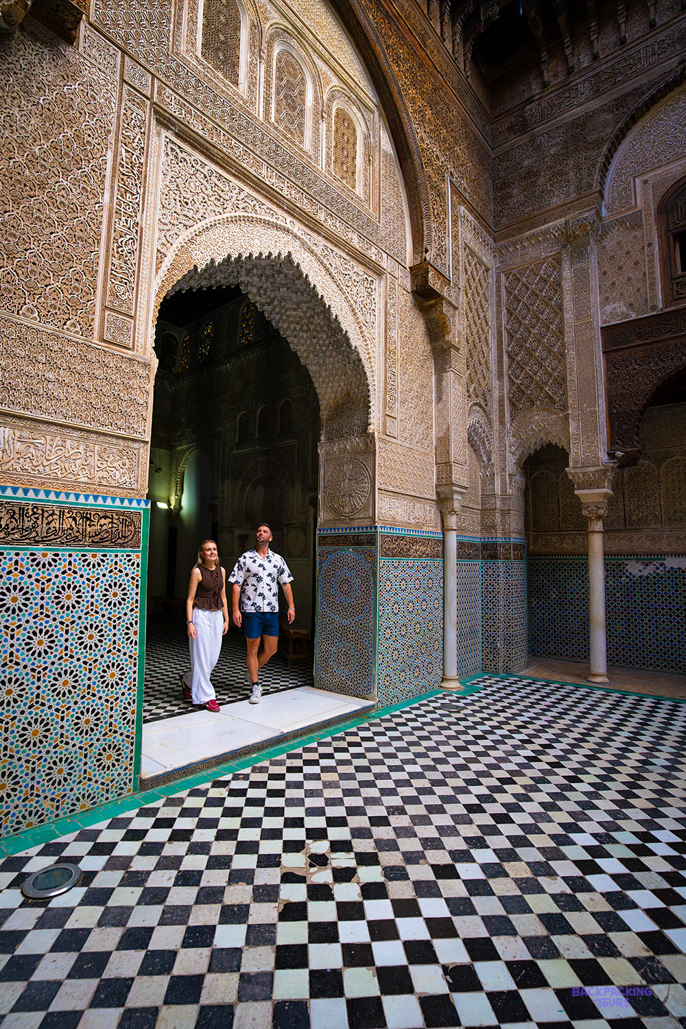 Two travellers stand in a carved stone doorway above a black and white chequered floor in Fes.