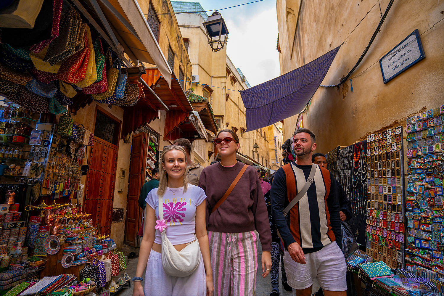 Four travellers walk down a narrow lane in the Fes medina lined with colourful shop displays.