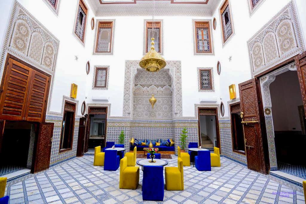 The tiled courtyard of Riad Dilar in Fes, with carved white walls and blue and yellow chairs.