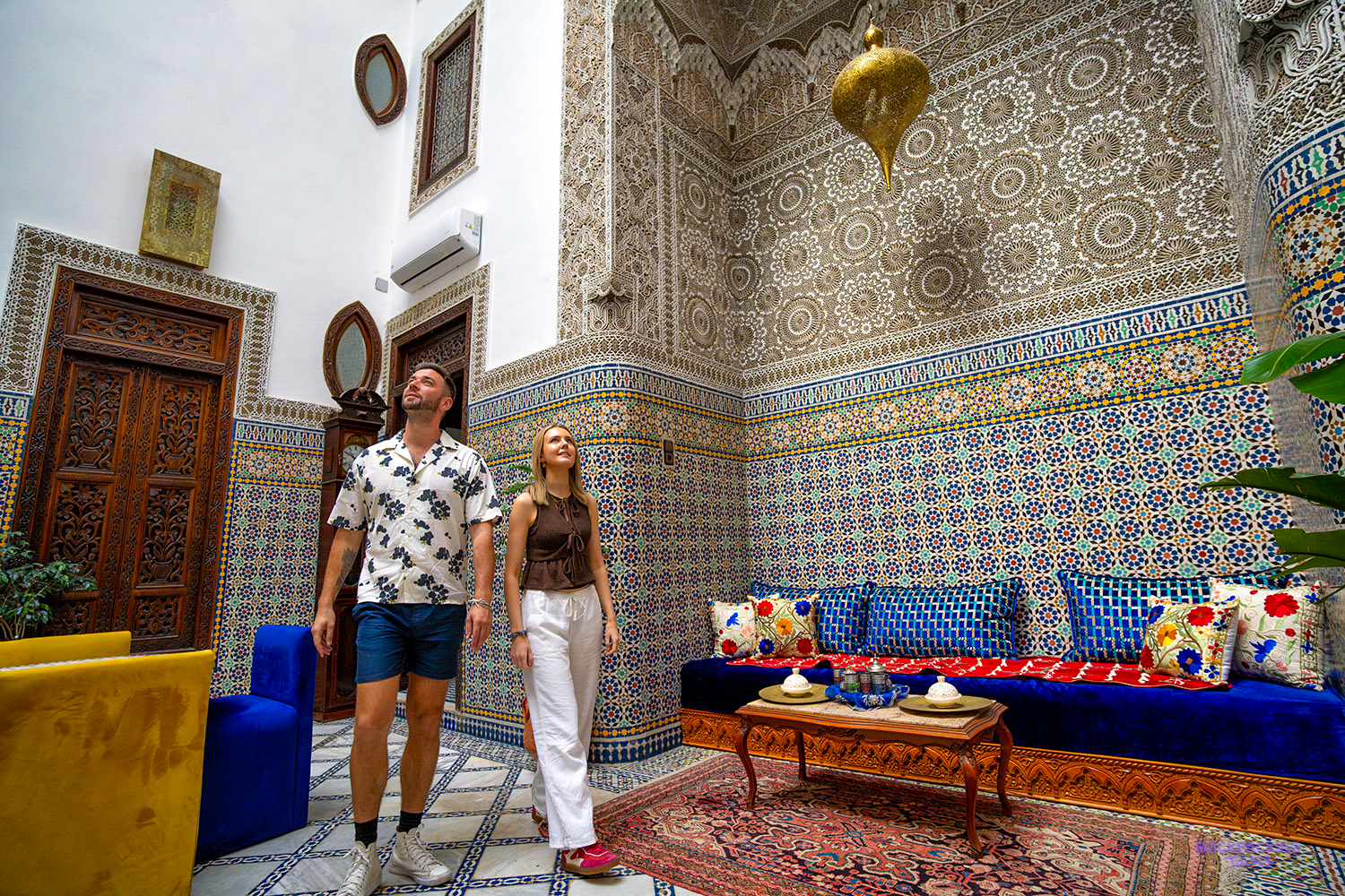 Two travellers in a Fes riad lounge with tiled walls, a blue sofa and a brass lantern.