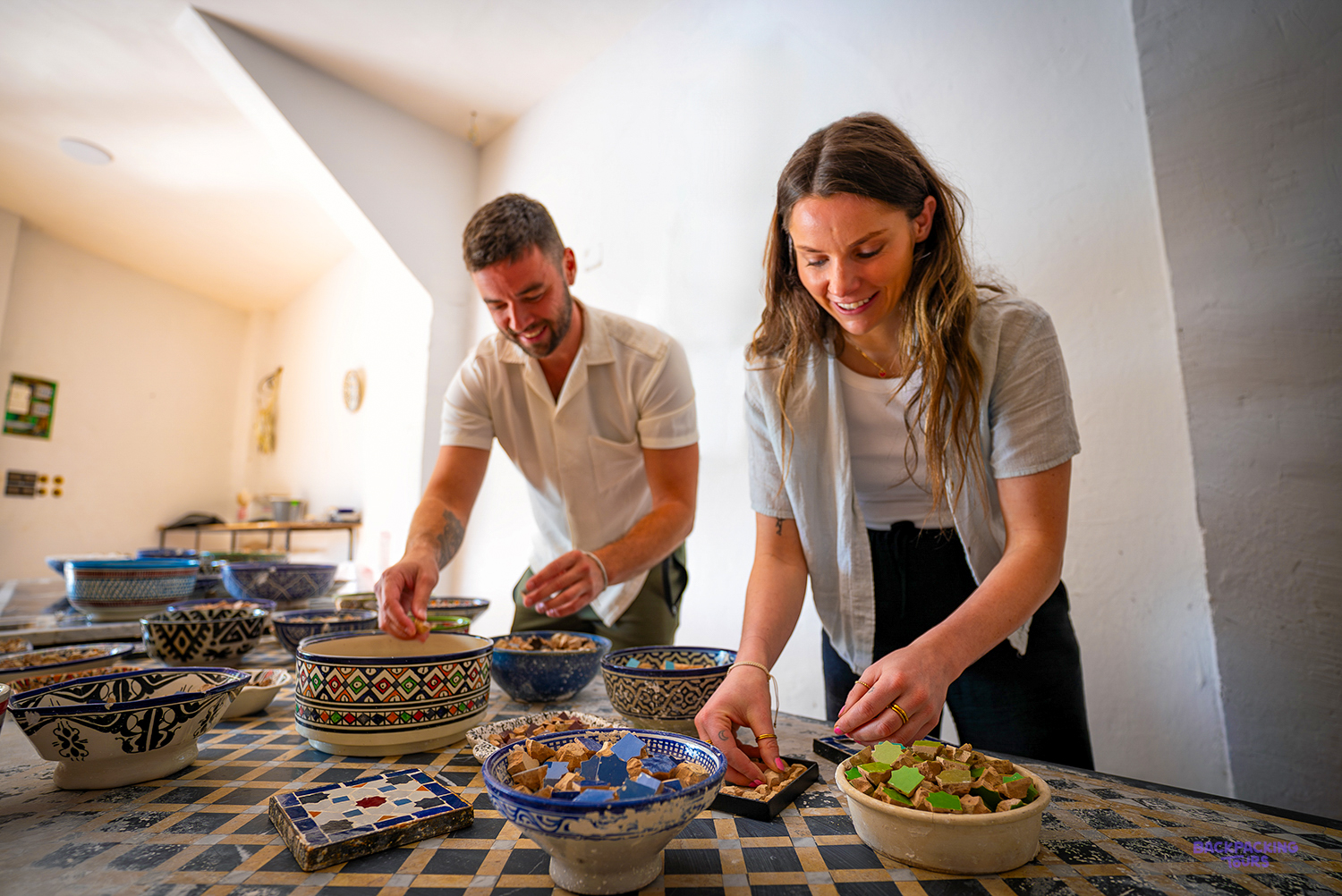 Two travellers arrange small cut tiles into patterns at a zellige mosaic workshop.