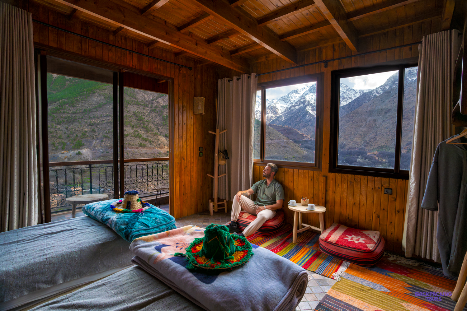 A traveller relaxes in a wood-panelled guesthouse room in Imlil, with snowy Atlas peaks through the windows.