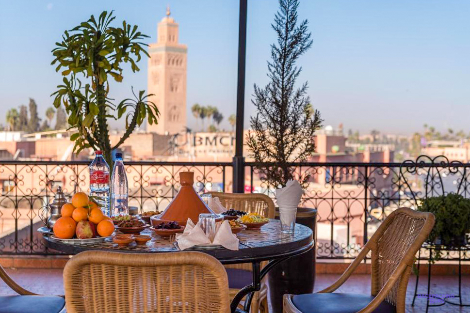 A table laid with a tagine and oranges on the rooftop terrace of Riad Omar, with a Marrakech minaret beyond.