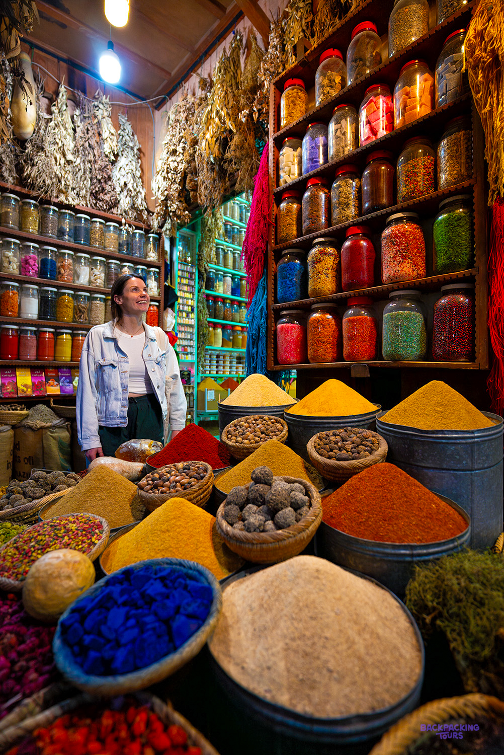 A traveller smiles beside jars of spices and cones of coloured powder at a Marrakech spice stall.
