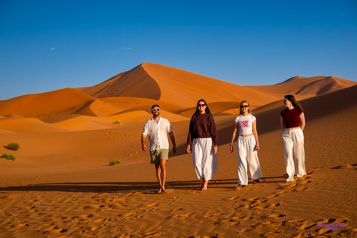 Four travellers walk across the sand towards the camera with a huge dune behind them.