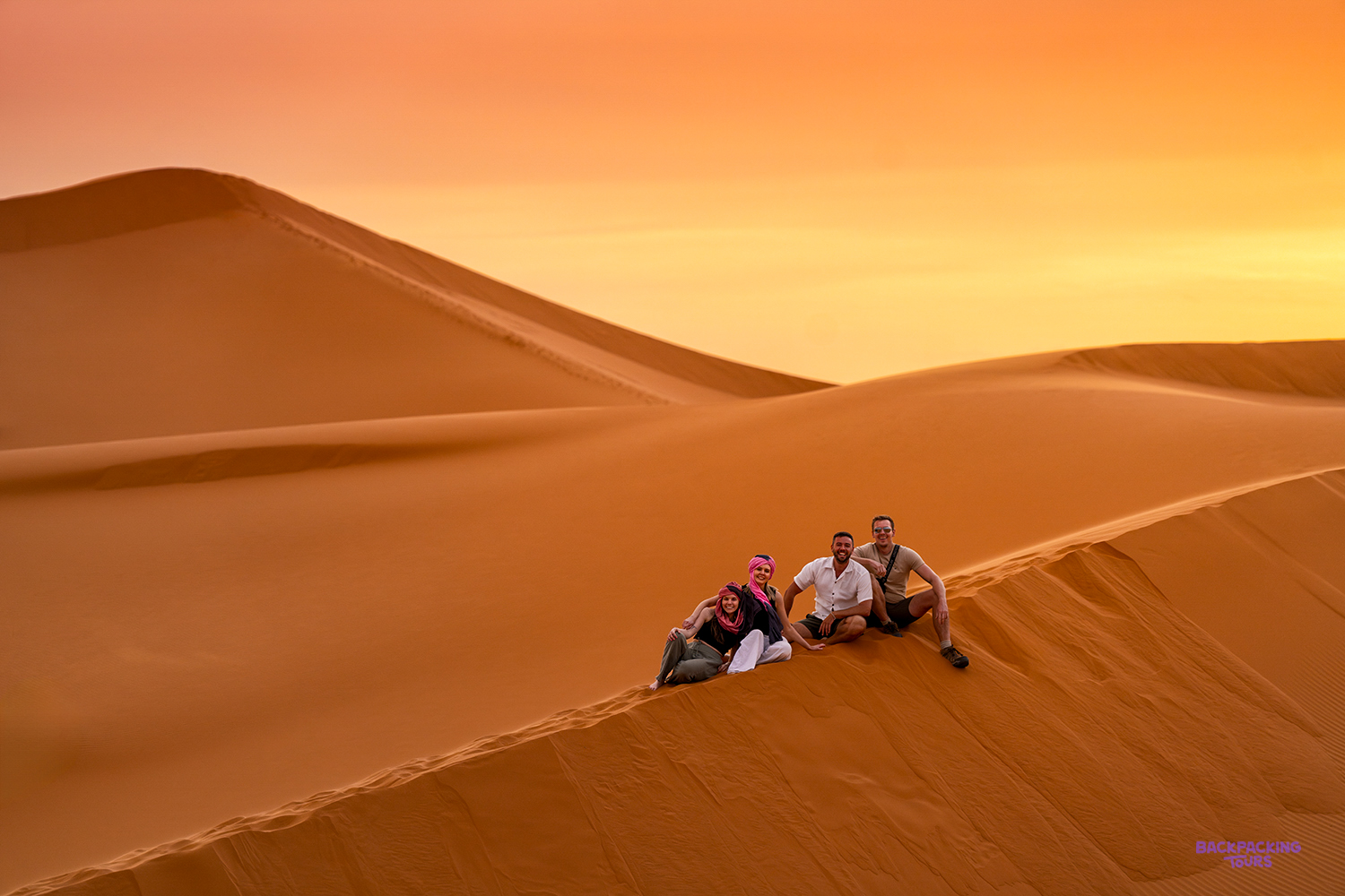 Travellers sit together on the crest of a Sahara dune in warm golden light.