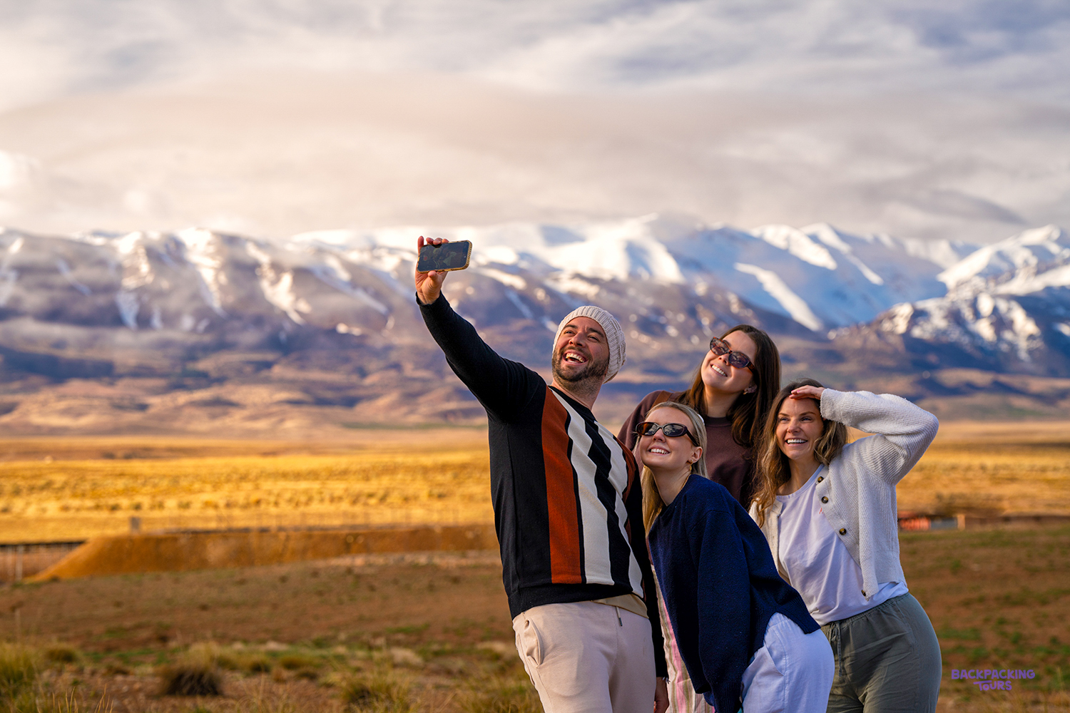Four travellers take a selfie on a golden plain with snow-capped Atlas mountains behind.