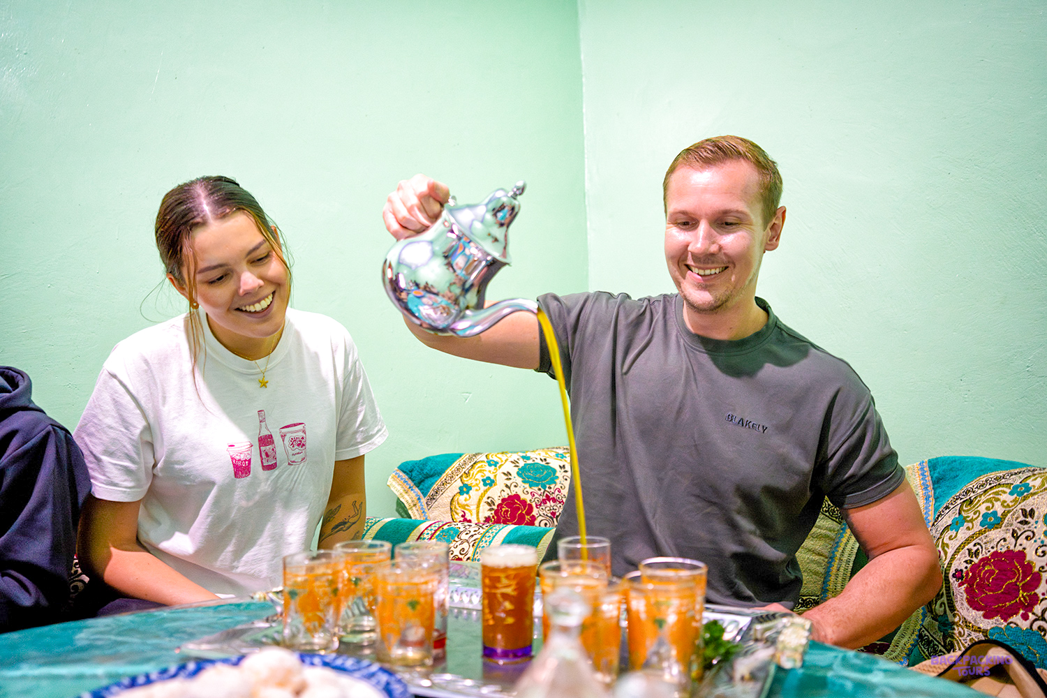 A smiling traveller pours mint tea from a silver teapot into small glasses at a family home.