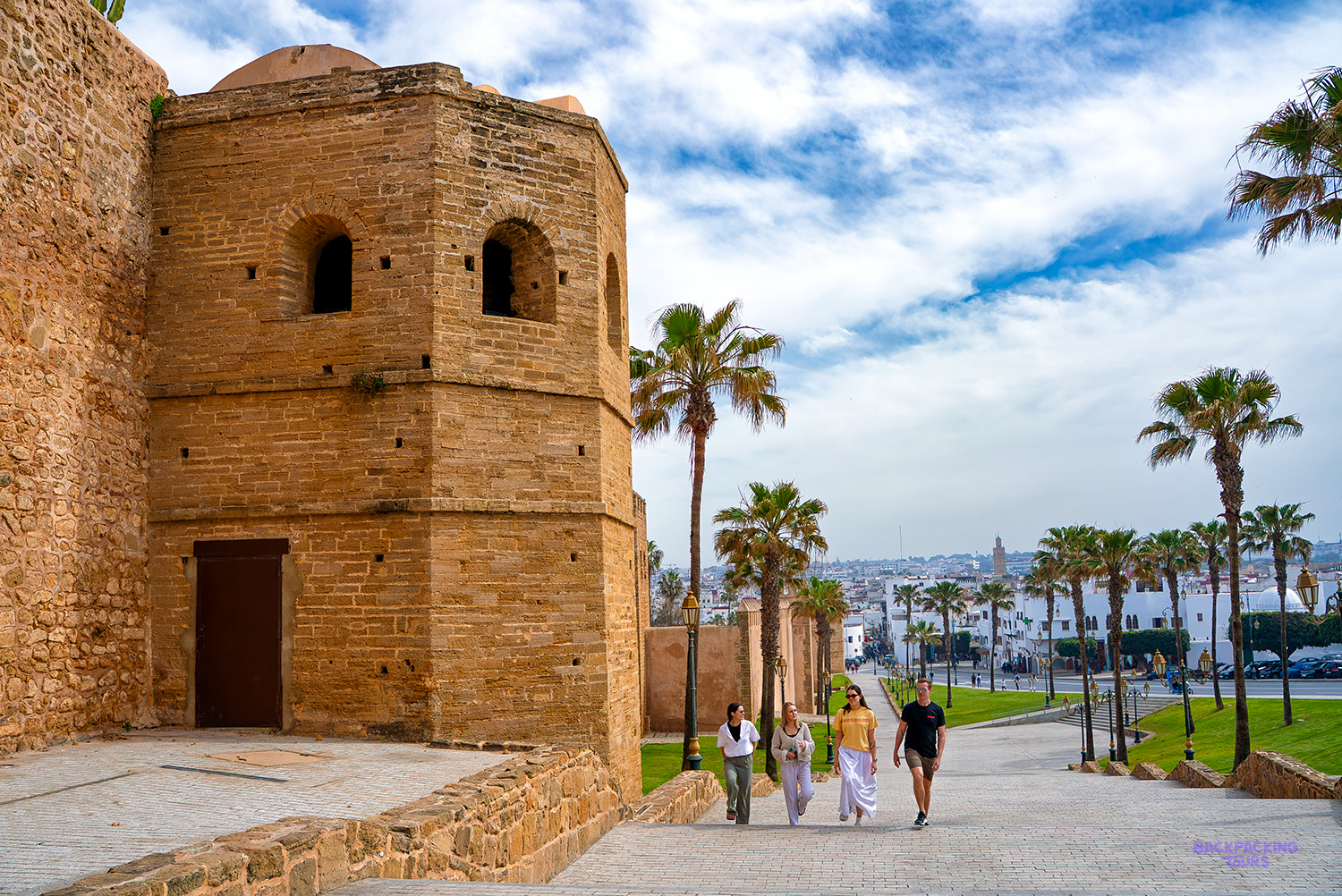 Travellers walk past an old stone tower and palm trees on a path beside Rabat's city walls.