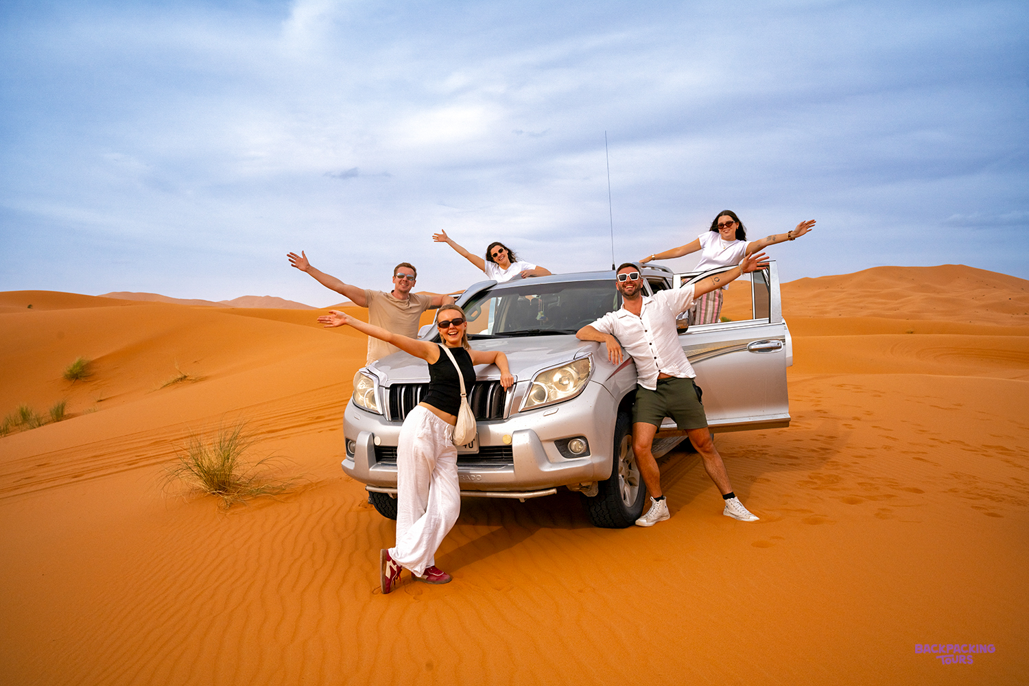 Travellers throw their arms out on and around a 4x4 parked on the Sahara dunes.