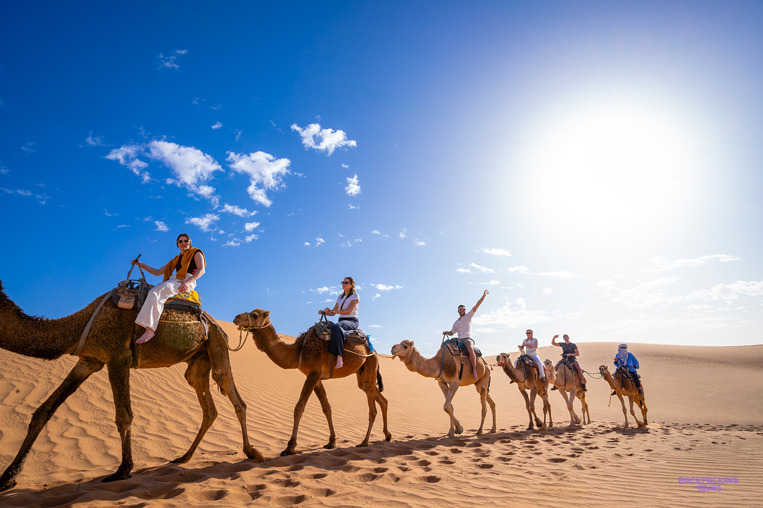 A line of travellers rides camels across the orange dunes of the Sahara on a sunny day.