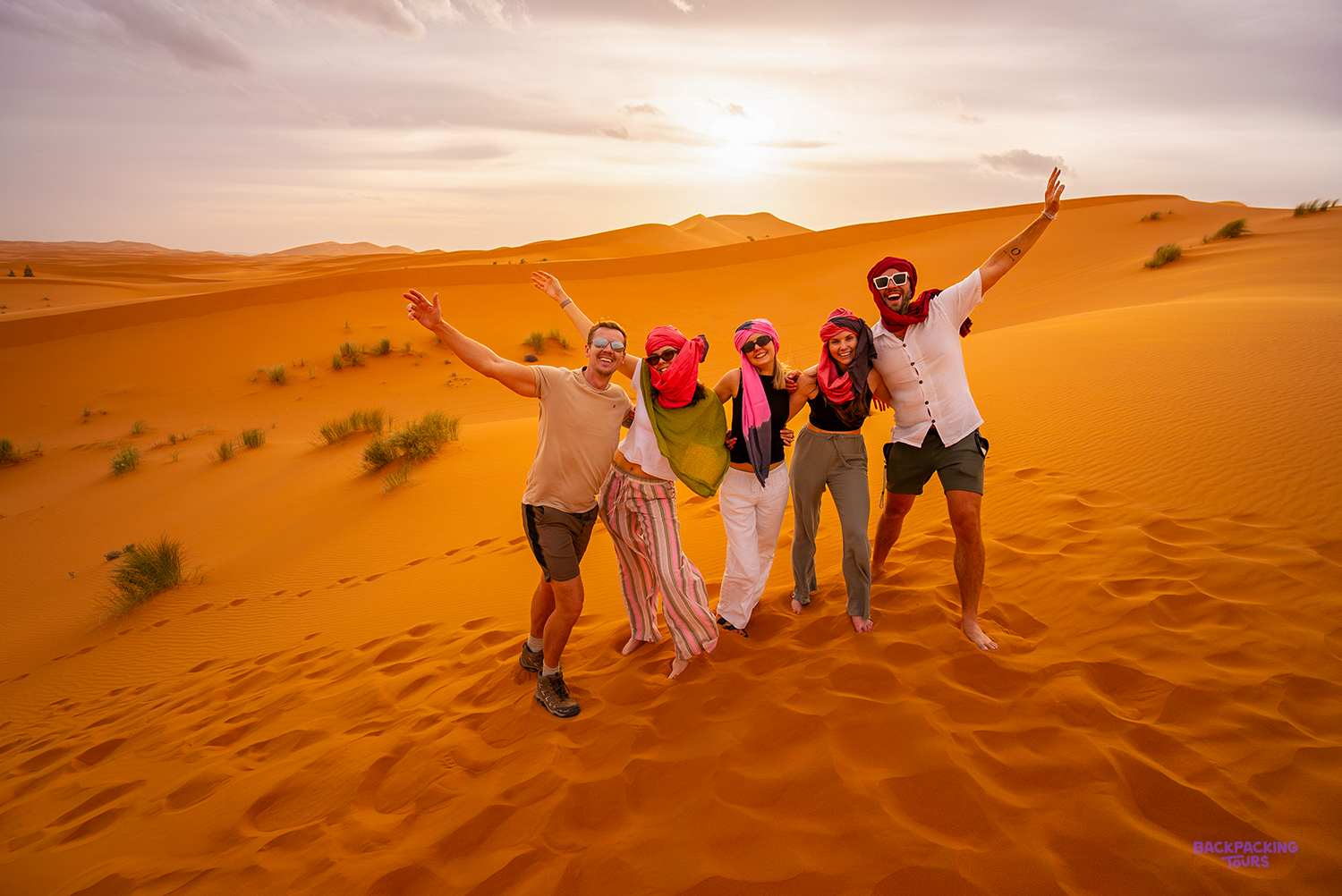 Travellers, some in colourful headscarves, cheer with raised arms on a Sahara dune in late afternoon light.