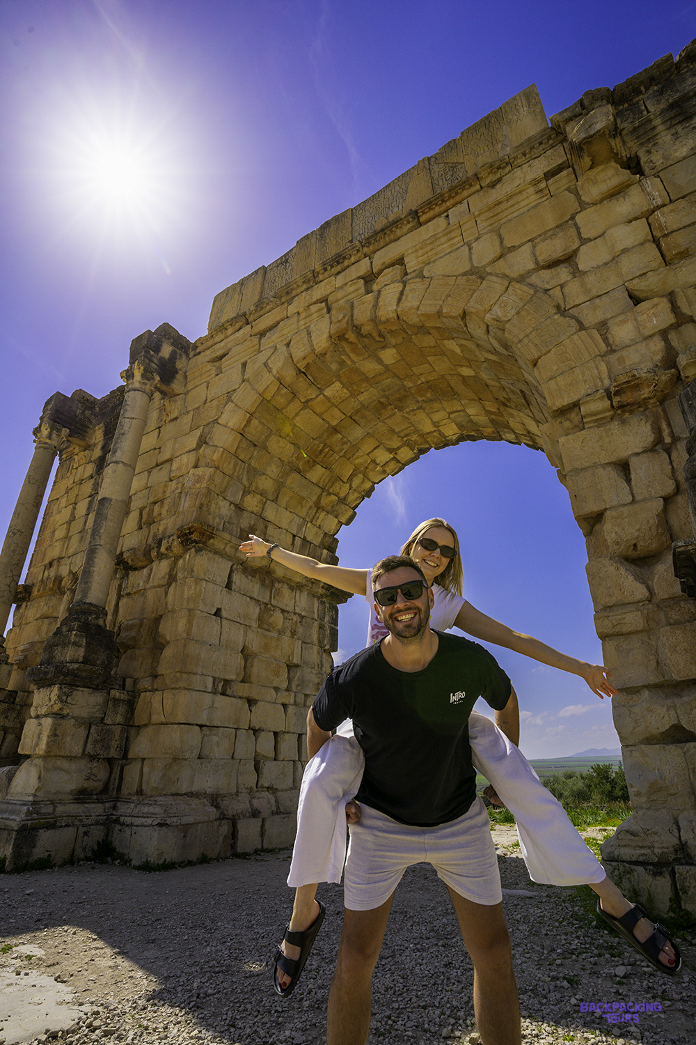 A traveller carries a friend on his back under a Roman stone arch at Volubilis.