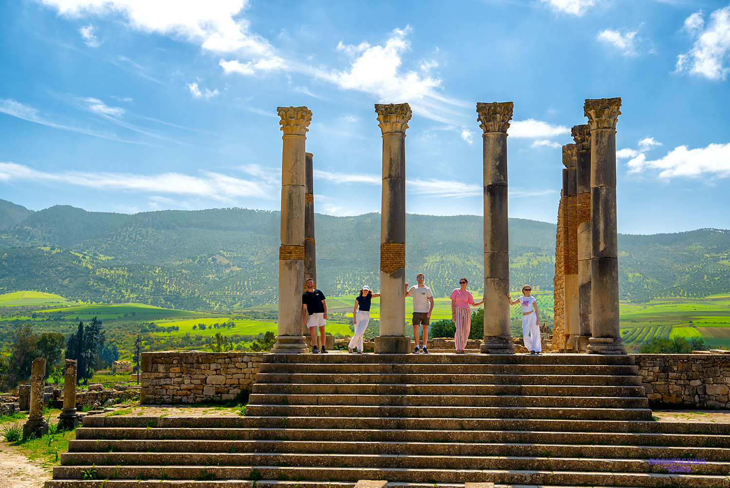Travellers stand between tall Roman columns at Volubilis, with green hills stretching behind them.