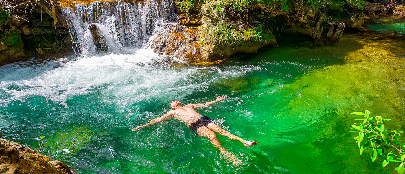 A swimmer floats in a clear green pool below a cascade at the Akchour Waterfalls.