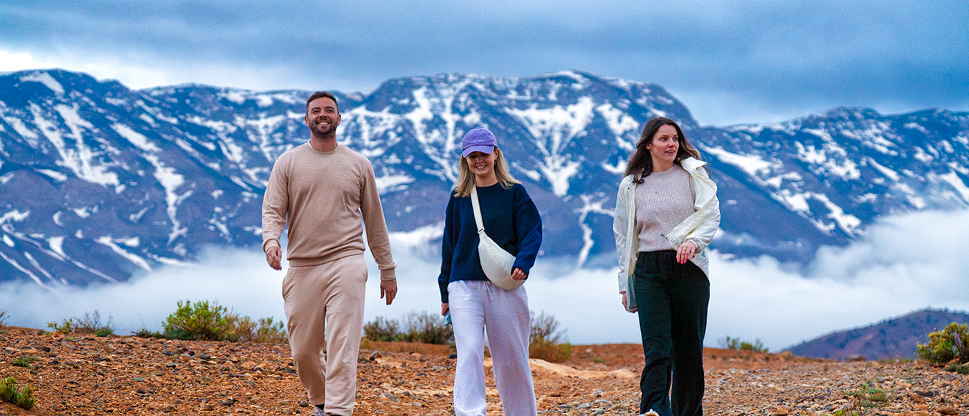 Three travellers walk along a mountain path above the clouds with snowy Atlas peaks behind.