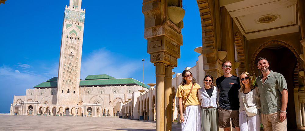 Five travellers smile beneath a carved archway with the tall minaret of the Hassan II Mosque behind.