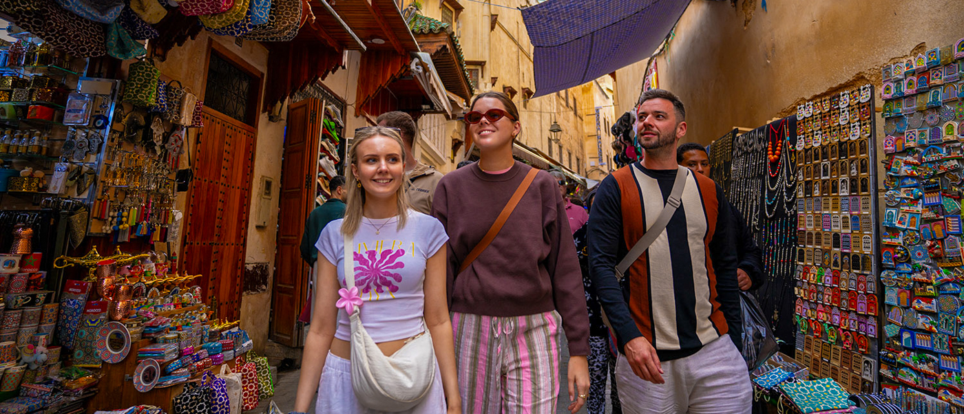 Four travellers walk down a narrow lane in the Fes medina lined with colourful shop displays.
