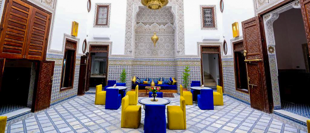 The tiled courtyard of Riad Dilar in Fes, with carved white walls and blue and yellow chairs.