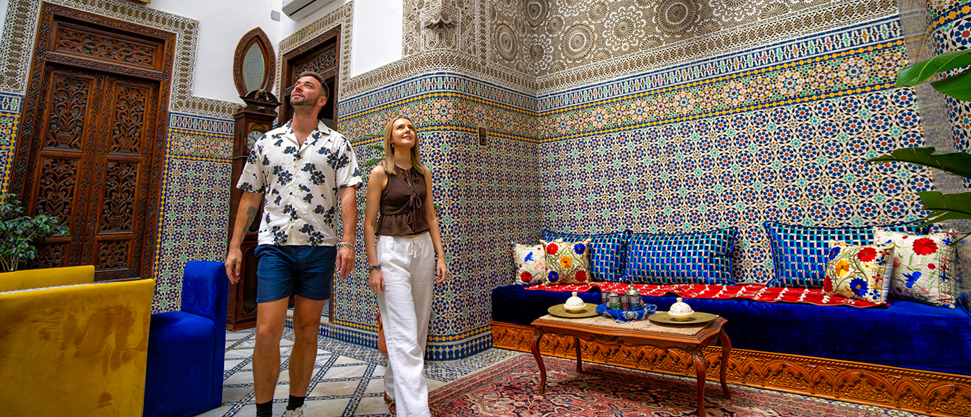 Two travellers in a Fes riad lounge with tiled walls, a blue sofa and a brass lantern.