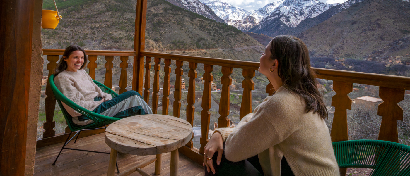 Two travellers relax on the wooden balcony of Dar Aymane Guesthouse facing snow-capped Atlas peaks.