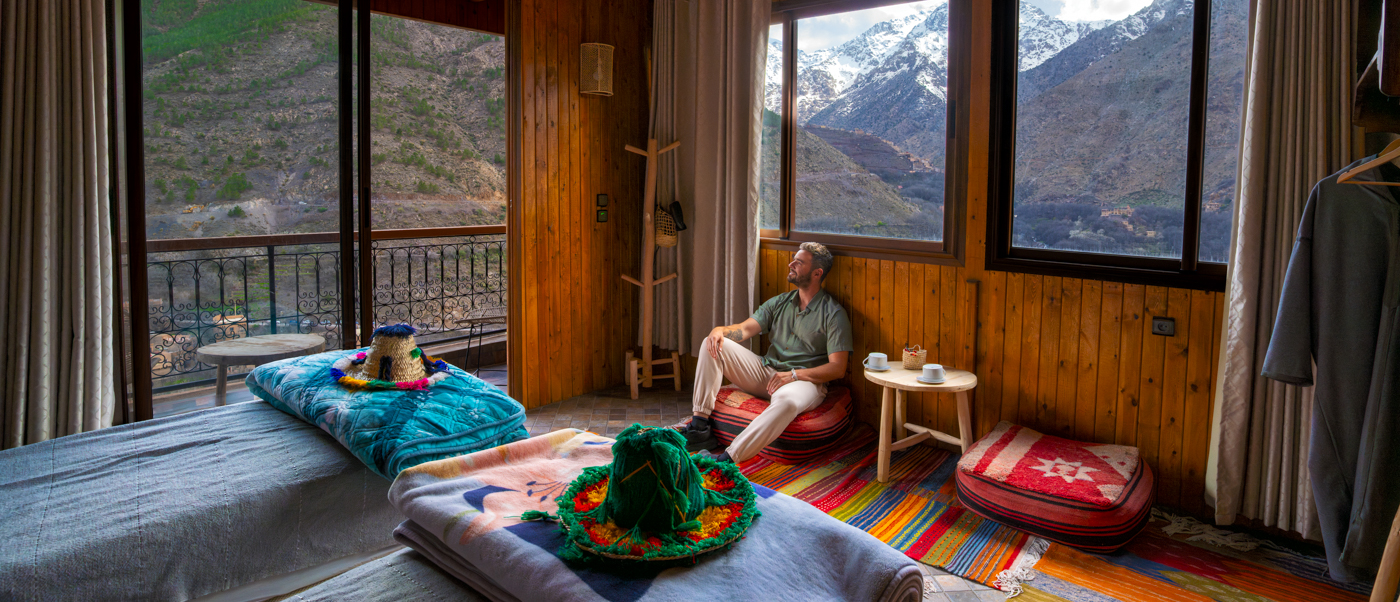 A traveller relaxes in a wood-panelled guesthouse room in Imlil, with snowy Atlas peaks through the windows.