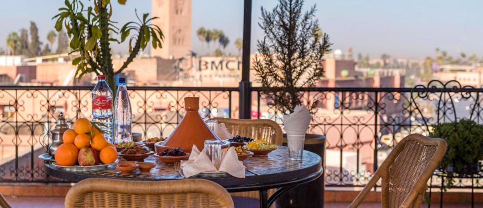 A table laid with a tagine and oranges on the rooftop terrace of Riad Omar, with a Marrakech minaret beyond.