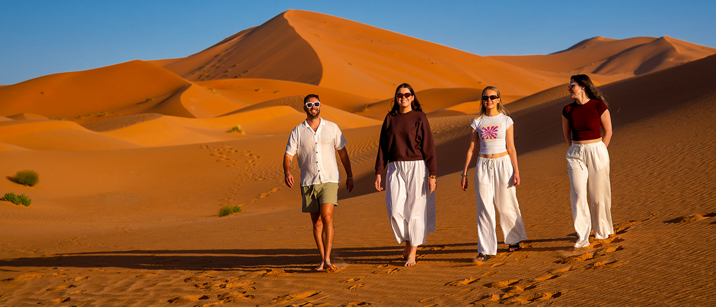 Four travellers walk across the sand towards the camera with a huge dune behind them.