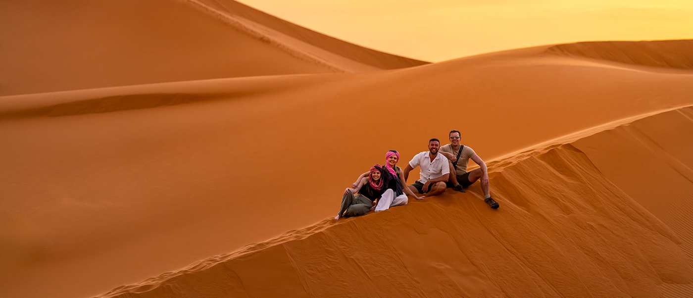 Travellers sit together on the crest of a Sahara dune in warm golden light.