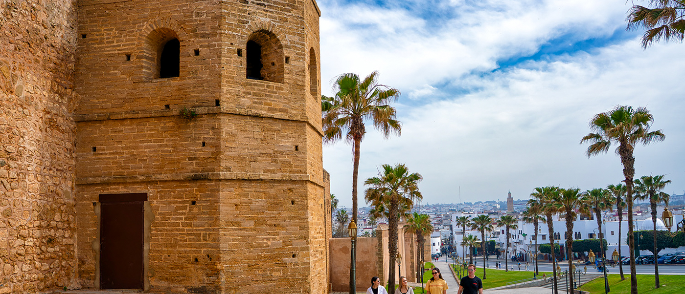 Travellers walk past an old stone tower and palm trees on a path beside Rabat's city walls.