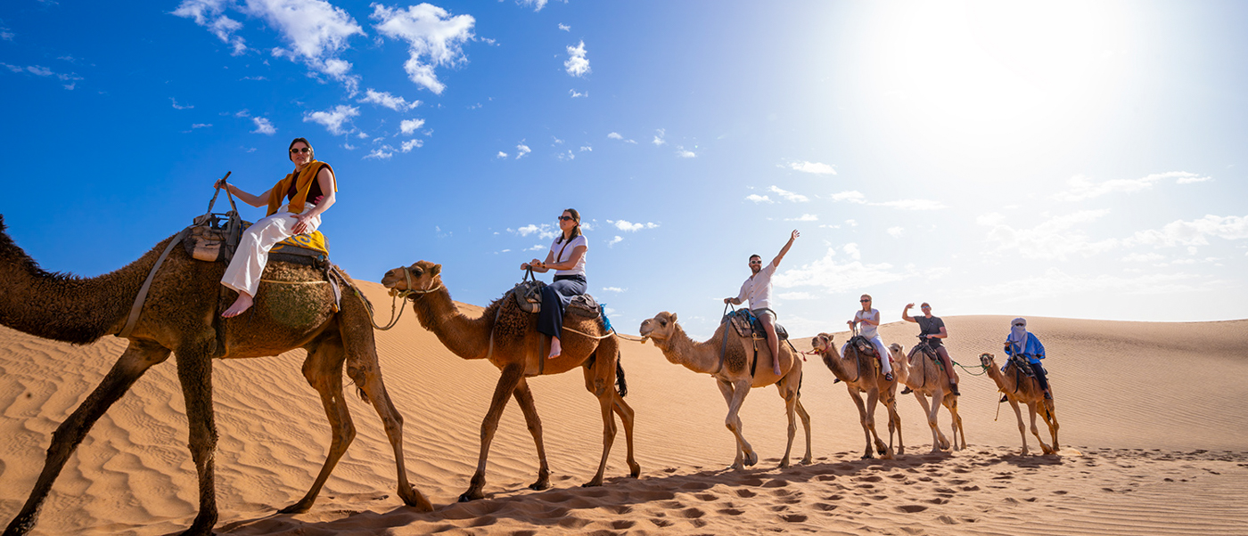 A line of travellers rides camels across the orange dunes of the Sahara on a sunny day.