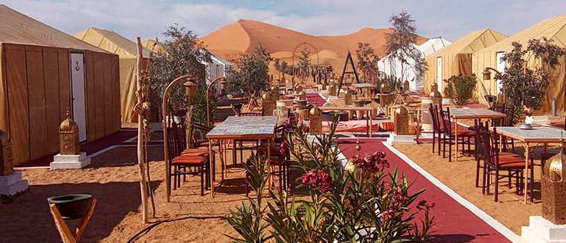 Rows of tents and dining tables at Tafouyte Camp, with a tall Sahara dune rising behind.