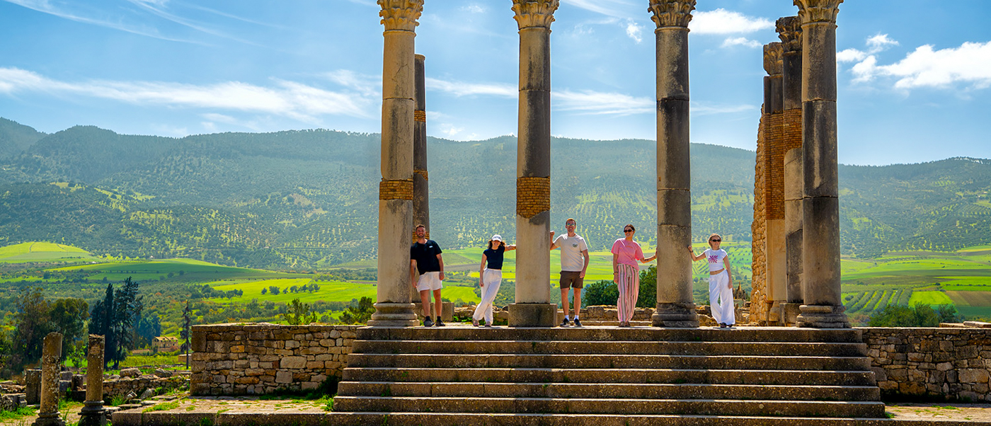 Travellers stand between tall Roman columns at Volubilis, with green hills stretching behind them.