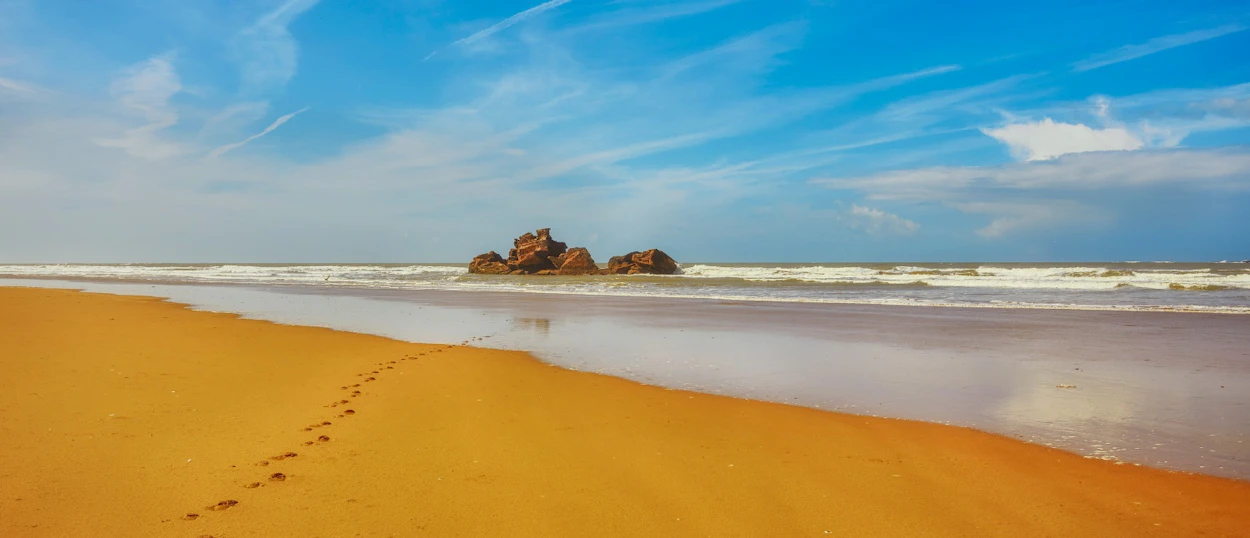 Essaouira beach with golden sand, footprints, ocean waves, and large rocks under a blue sky.