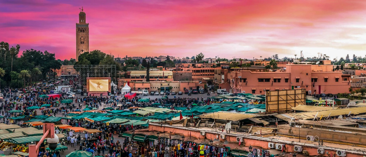 A vibrant market square in Marrakech at sunset, with a tall minaret and many bustling stalls.