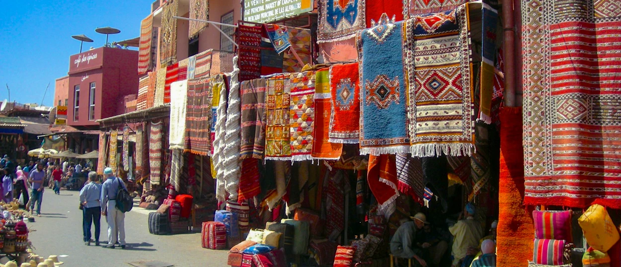 People browse colorful rugs and textiles hanging from stalls in a bustling Marrakech market.