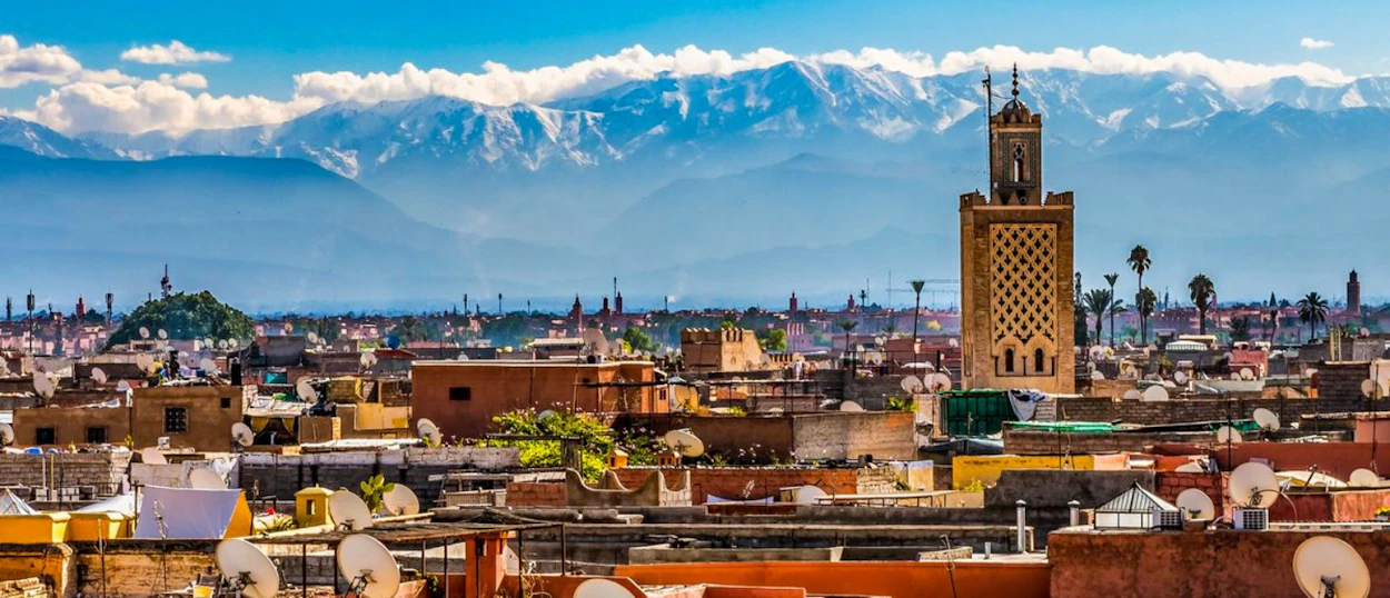 Rooftops of Marrakech with a tall minaret under a blue sky and distant snow-capped Atlas Mountains.