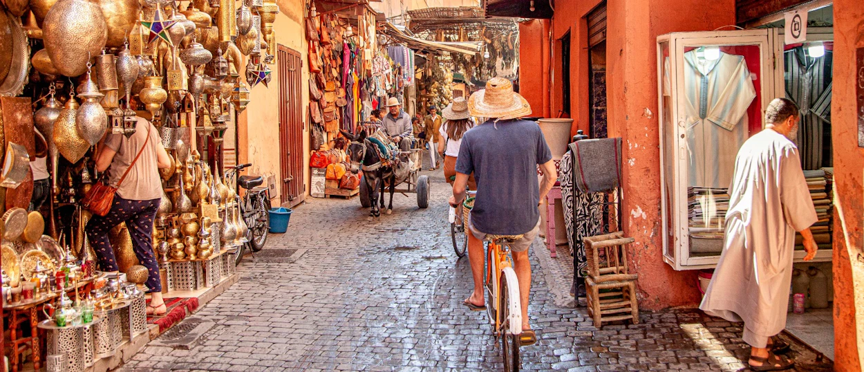 People browse and cycle through a bustling Marrakech souk filled with traditional lamps and goods.