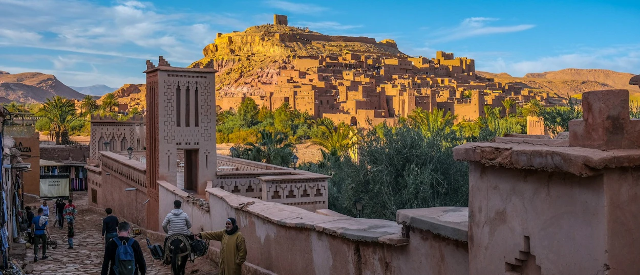 Backpackers explore the historic Ait Ben Haddou kasbah, a fortified village nestled against a sunlit hill.